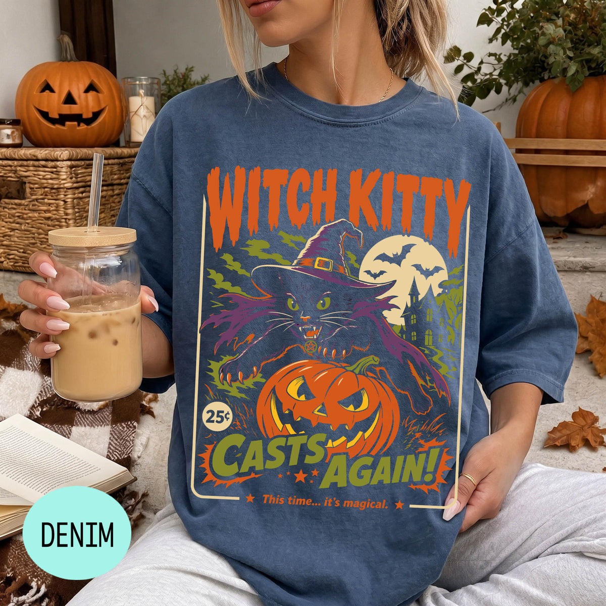 A woman wearing a blue t-shirt with a Halloween-themed graphic, holding a drink and surrounded by pumpkins and other fall decorations.