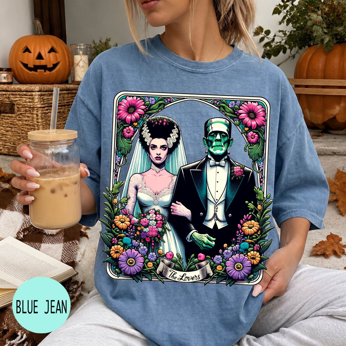 A woman wearing a blue t-shirt with a graphic design featuring a bride and a Frankenstein monster, holding a drink and surrounded by Halloween decorations.