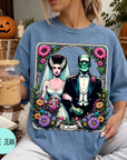 A woman wearing a blue t-shirt with a graphic design featuring a bride and a Frankenstein monster, holding a drink and surrounded by Halloween decorations.