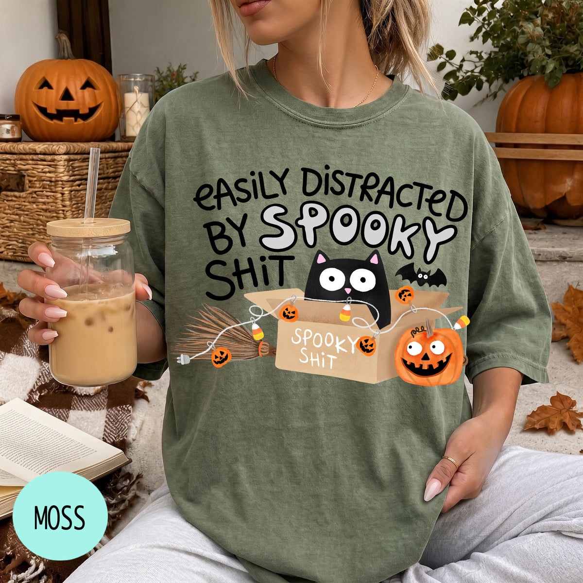 A person wearing a green t-shirt with a humorous Halloween-themed graphic and text, holding a drink and surrounded by Halloween decorations.