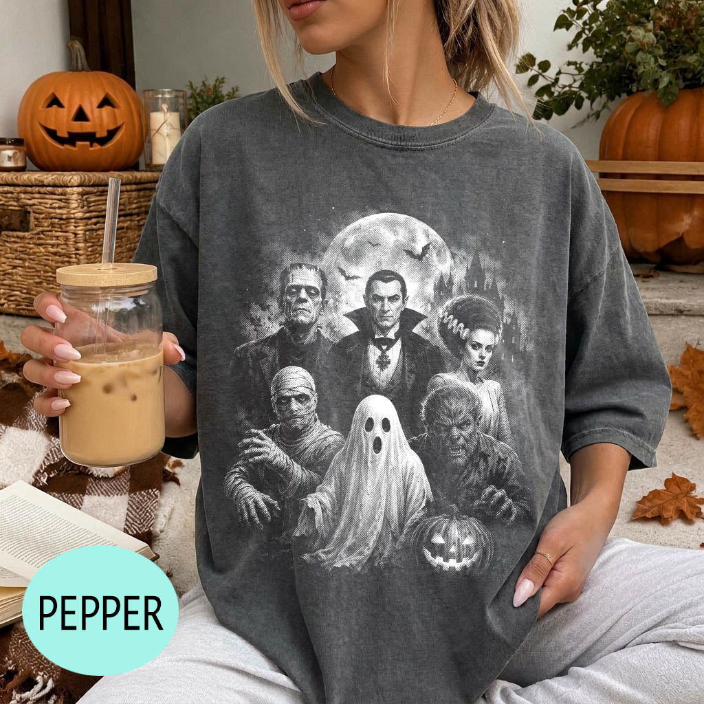 A woman wearing a t-shirt with a Halloween-themed graphic of the classic horror movie characters, including Frankenstein, Dracula, Frankenstein's monster, Frankenstein's ghost, and the werewolf. She is holding a drink in a glass with a straw.