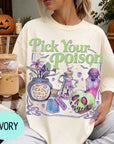 A woman wearing a white t-shirt with a graphic design featuring various Halloween-themed items, such as a skull, a vase, and a bottle, along with the text "Pick Your Poison". She is holding a drink in a glass with a straw.