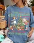 A woman wearing a blue t-shirt with a graphic design of a Christmas tree and two children decorating it, holding a drink and sitting on a blanket.