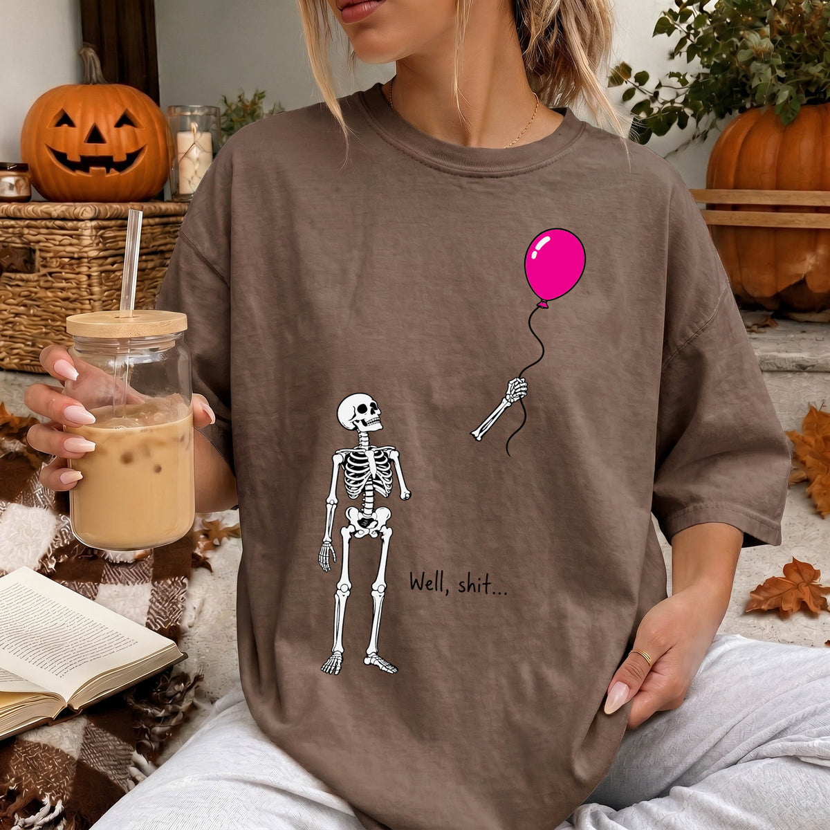 A woman wearing a t-shirt with a skeleton and a balloon graphic, holding a glass of iced coffee and a book.