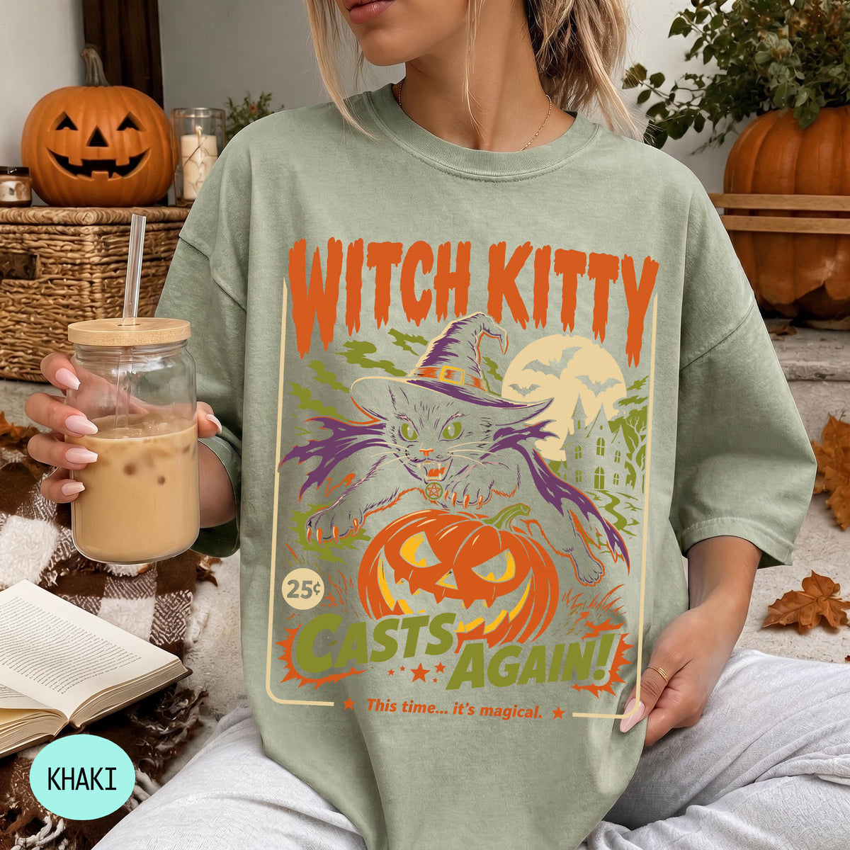 A woman wearing a green t-shirt with a Halloween-themed graphic design, including a witch, pumpkin, and text, is holding a drink in her hand.