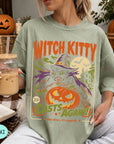 A woman wearing a green t-shirt with a Halloween-themed graphic design, including a witch, pumpkin, and text, is holding a drink in her hand.