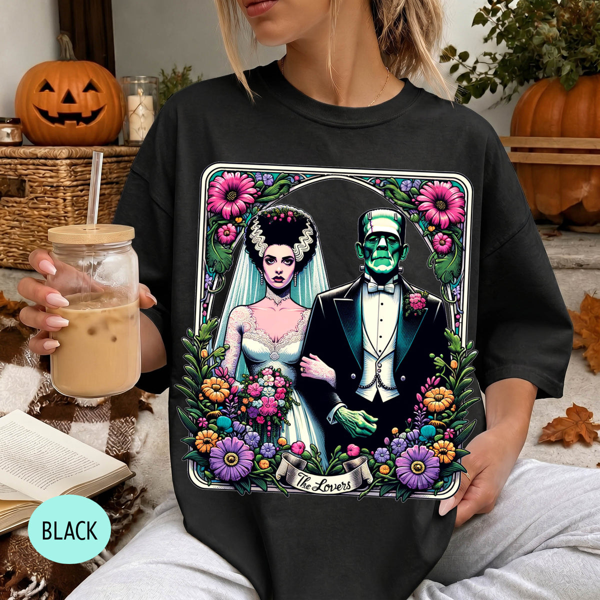A woman wearing a black t-shirt with a graphic design featuring a bride and a Frankenstein monster, surrounded by flowers and a banner that says "The Lovers".