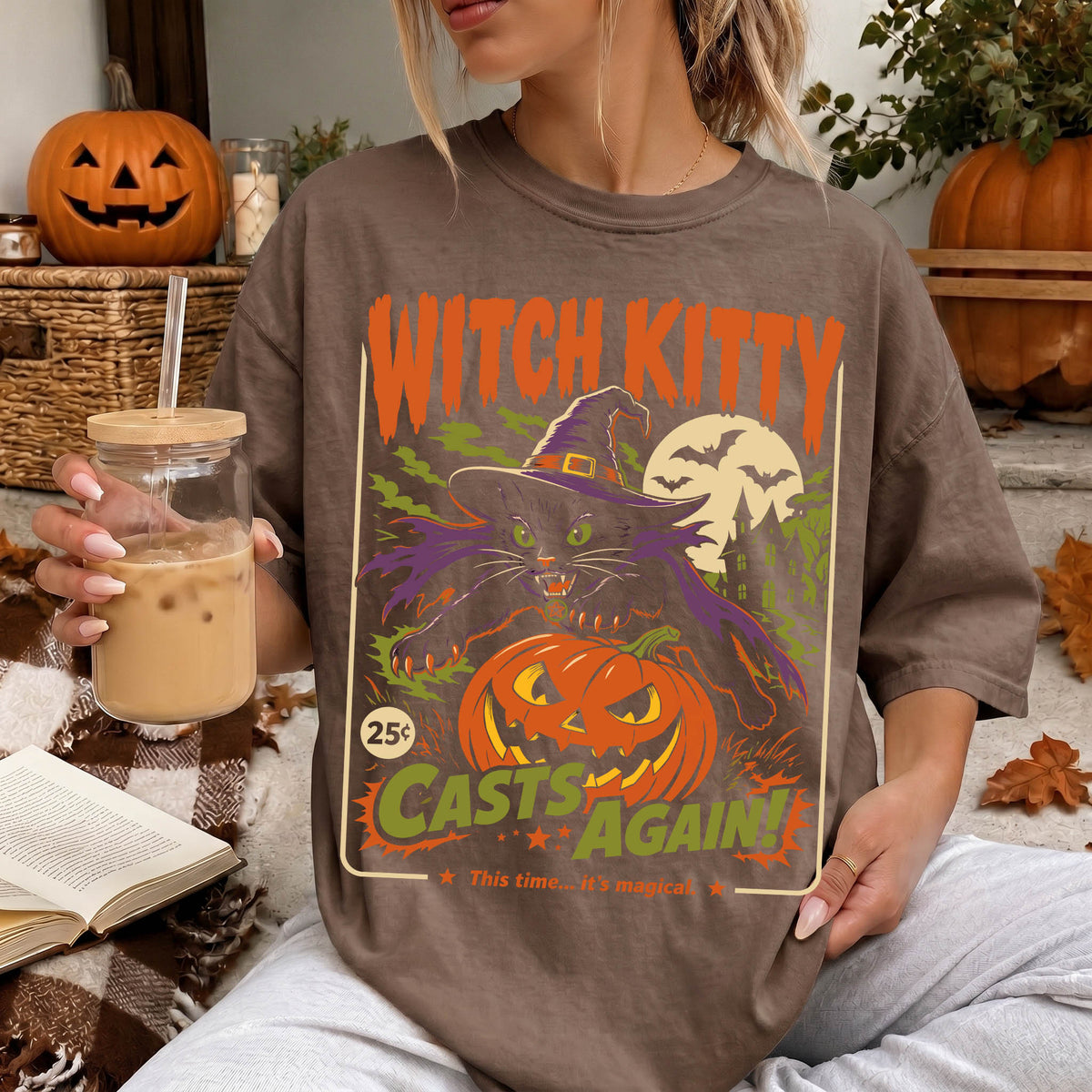 A woman wearing a t-shirt with a Halloween-themed graphic, holding a drink and surrounded by pumpkins and autumn leaves.