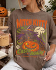 A woman wearing a t-shirt with a Halloween-themed graphic, holding a drink and surrounded by pumpkins and autumn leaves.