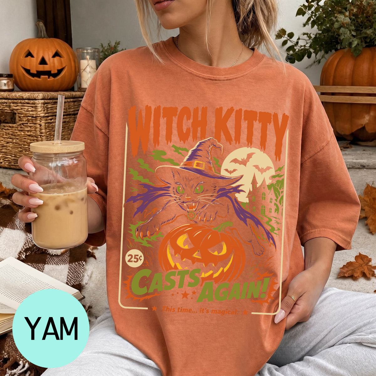 A woman wearing a t-shirt with a Halloween-themed design, including a witch, pumpkin, and bats, is holding a drink in her hand.