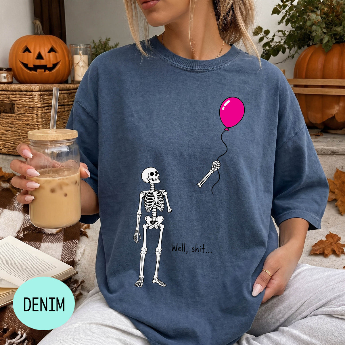 A woman wearing a blue t-shirt with a skeleton and a balloon graphic, holding a drink and sitting on a blanket with a pumpkin and autumn leaves in the background.