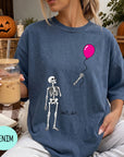 A woman wearing a blue t-shirt with a skeleton and a balloon graphic, holding a drink and sitting on a blanket with a pumpkin and autumn leaves in the background.