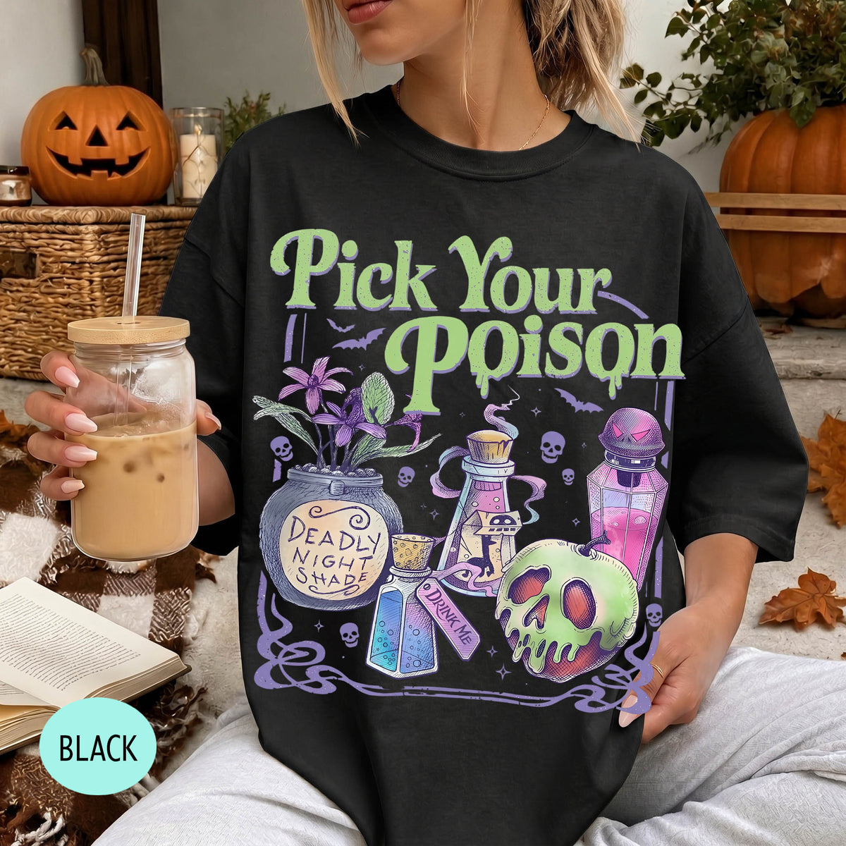 A woman wearing a black t-shirt with a Halloween-themed graphic design, holding a drink and surrounded by Halloween decorations.