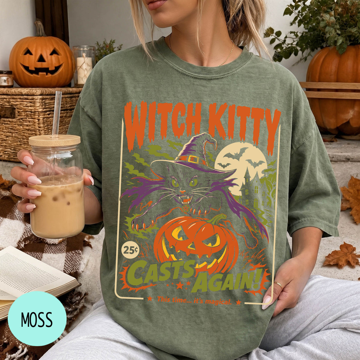 A woman wearing a green t-shirt with a Halloween-themed graphic and holding a drink, with a pumpkin and other fall decorations visible in the background.