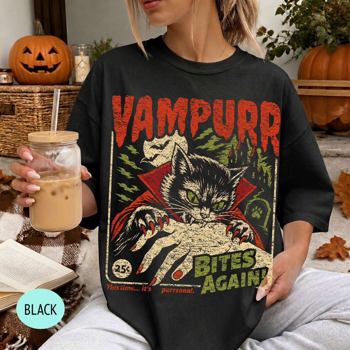 A woman wearing a black t-shirt with a graphic design of a cat wearing a cape and holding a bat, with the text "VAMPURR BITE'S AGAIN" printed on it. She is holding a drink in a glass with a straw.