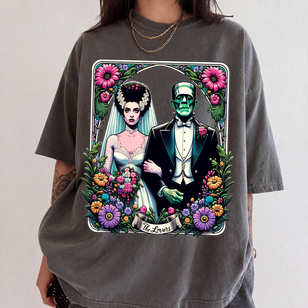 A graphic t-shirt featuring a bride and groom with a greenish-blue monster figure, surrounded by flowers and the text "The Lovers".