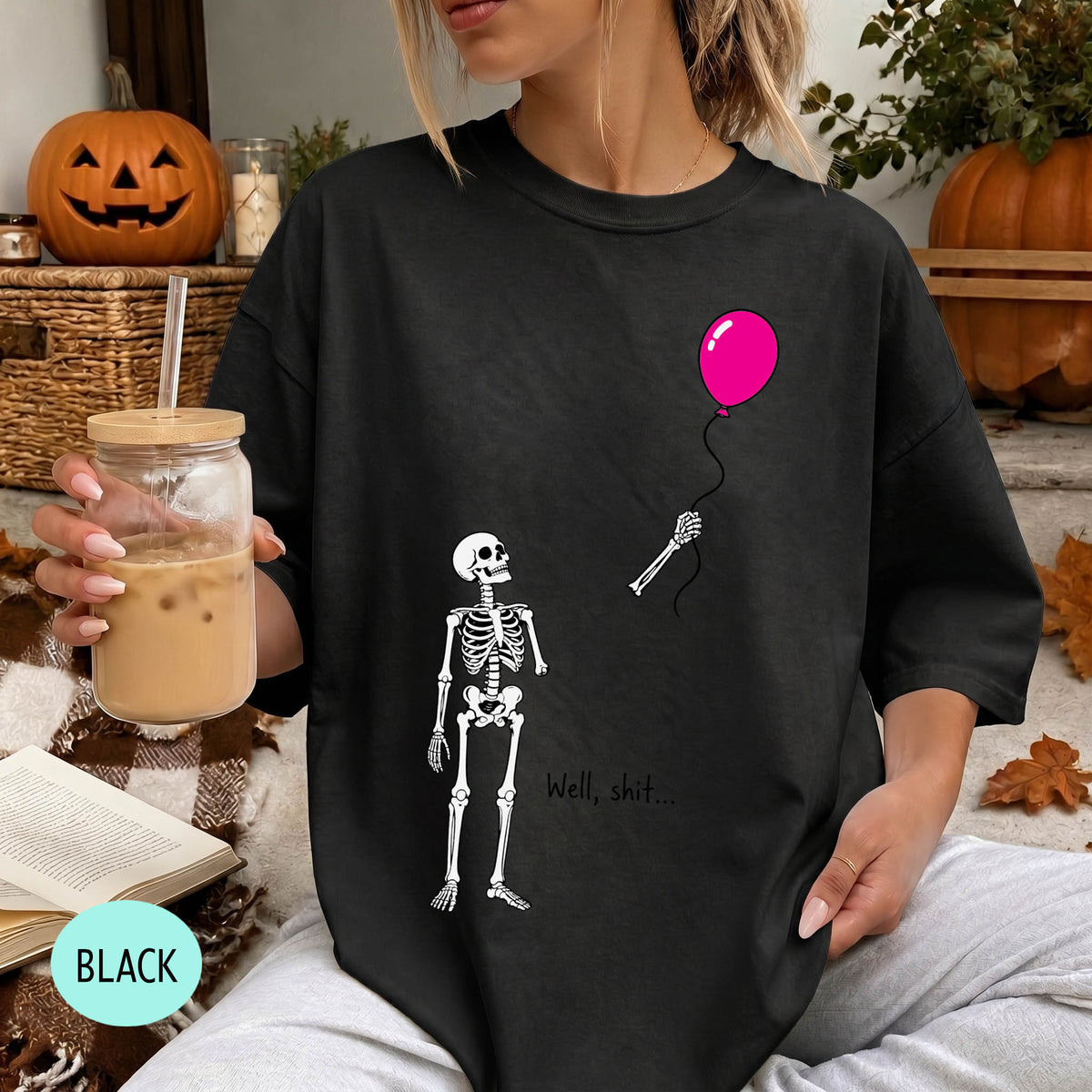 A woman wearing a black t-shirt with a skeleton and a pink balloon graphic, holding a drink and sitting on a blanket with a pumpkin and autumn leaves in the background.