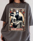 a graphic t-shirt with a design depicting a woman in a gown and a man in a suit, surrounded by various symbols and text, including the words "MONSTER", "THE LOVERS", and "THE LOVERS".