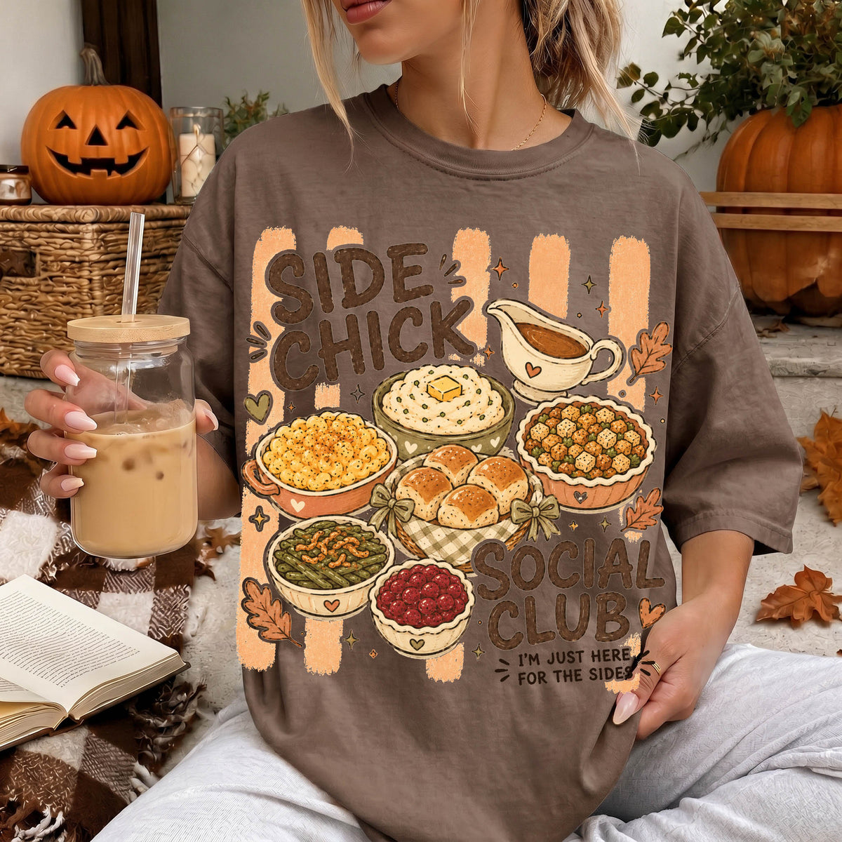 A woman wearing a t-shirt with a graphic design of various dishes and a pumpkin, holding a drink and surrounded by fall decorations.