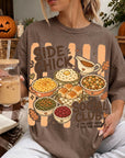 A woman wearing a t-shirt with a graphic design of various dishes and a pumpkin, holding a drink and surrounded by fall decorations.