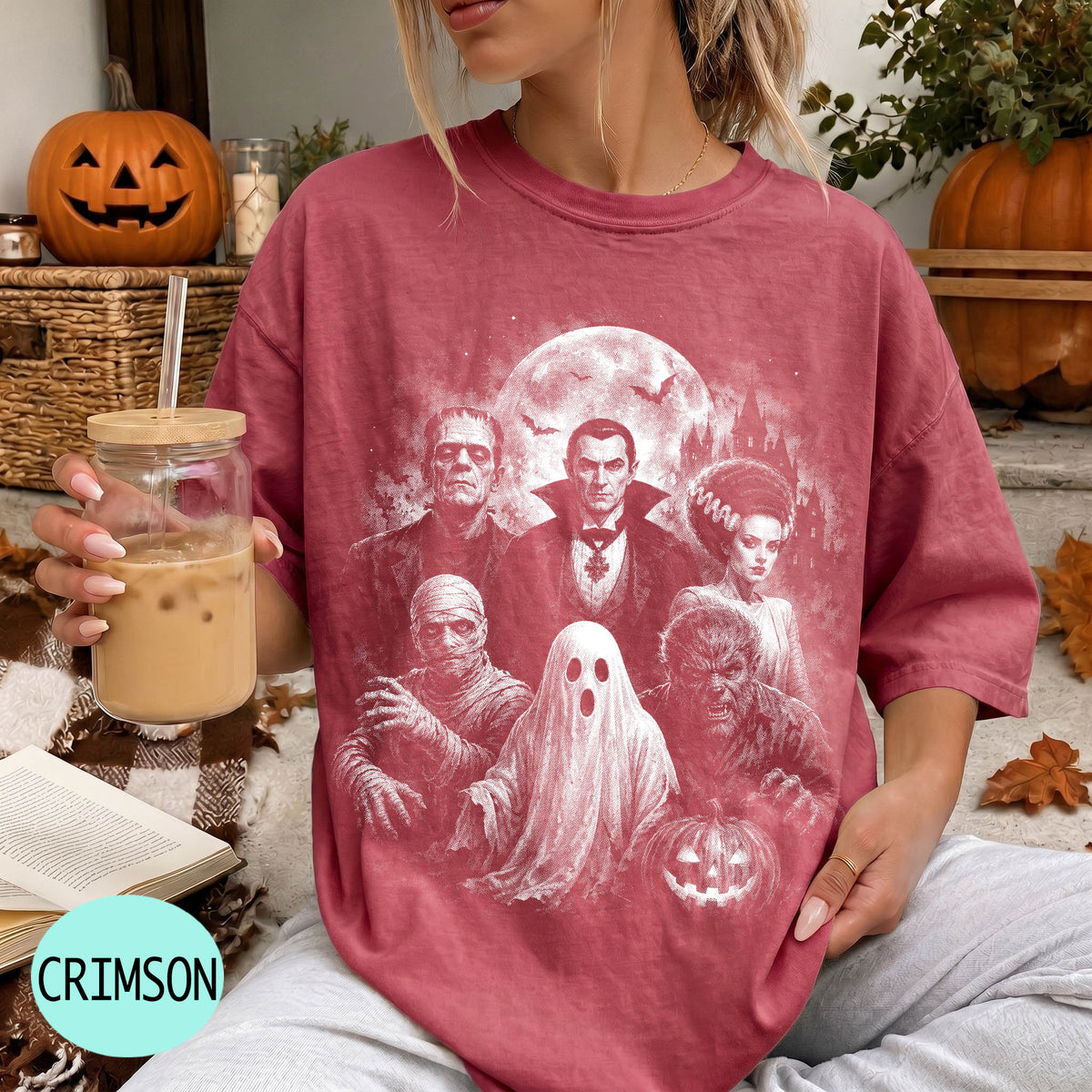 A woman wearing a red t-shirt with a Halloween-themed graphic of Dracula, Frankenstein, Frankenstein's Bride, Frankenstein's monster, and a ghost, holding a drink and sitting on a blanket.