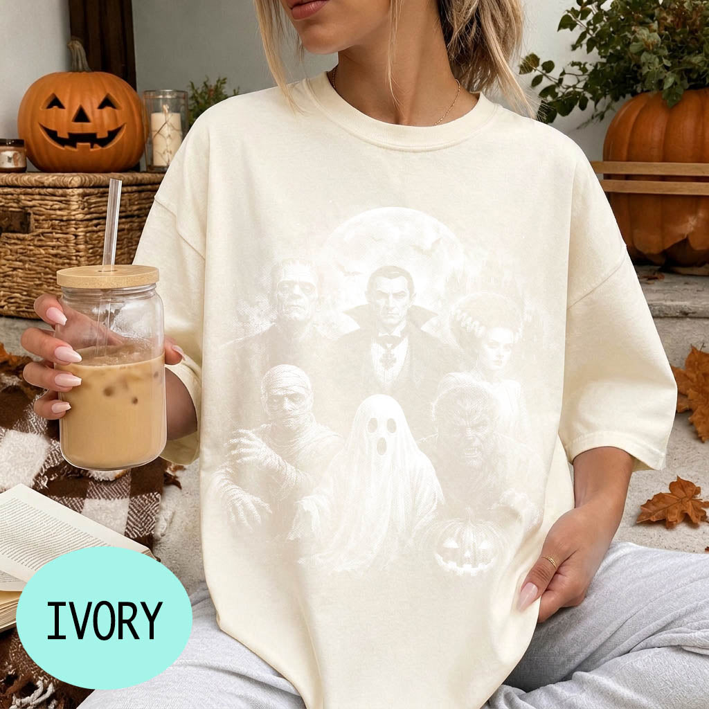 A woman wearing a white t-shirt with a Halloween-themed graphic, holding a glass of iced coffee, and surrounded by Halloween decorations.