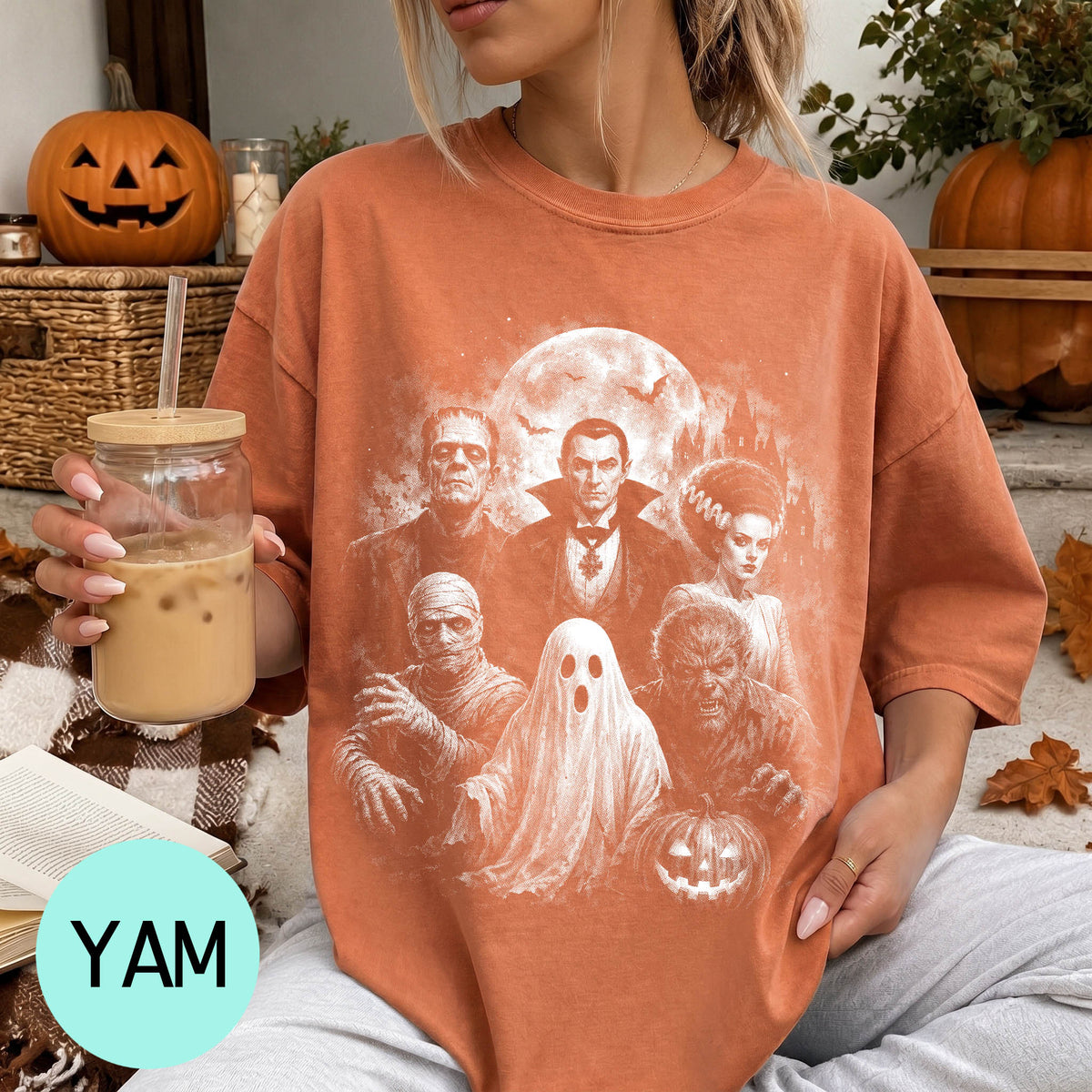 A woman wearing a t-shirt with a Halloween-themed graphic of characters from the horror film "The Bride of Frankenstein" is holding a drink and sitting on a couch with a pumpkin and other Halloween decorations in the background.