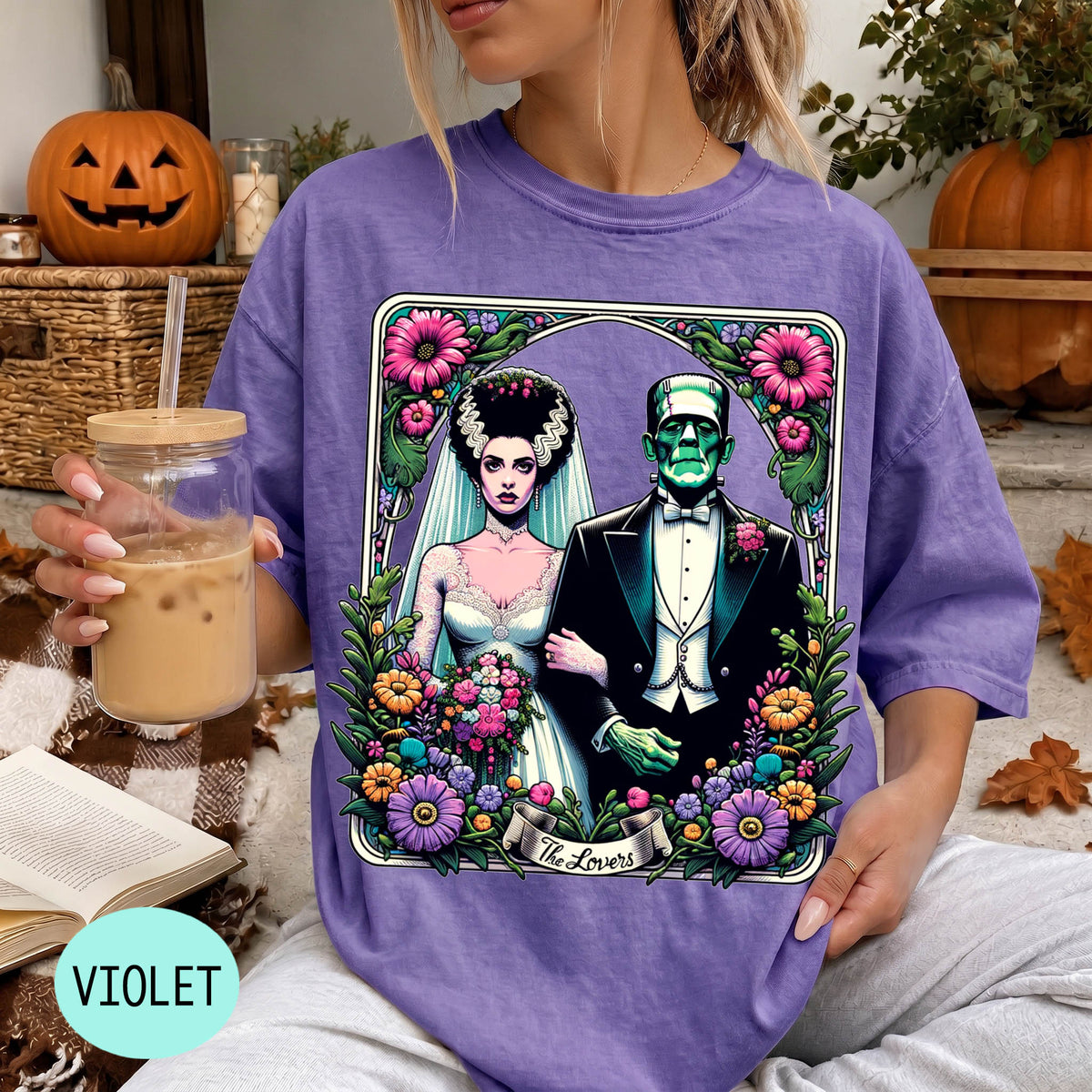 A woman wearing a purple t-shirt with a graphic design featuring a bride and groom, surrounded by flowers and a banner that says "The Lovers". She is holding a drink in a glass.
