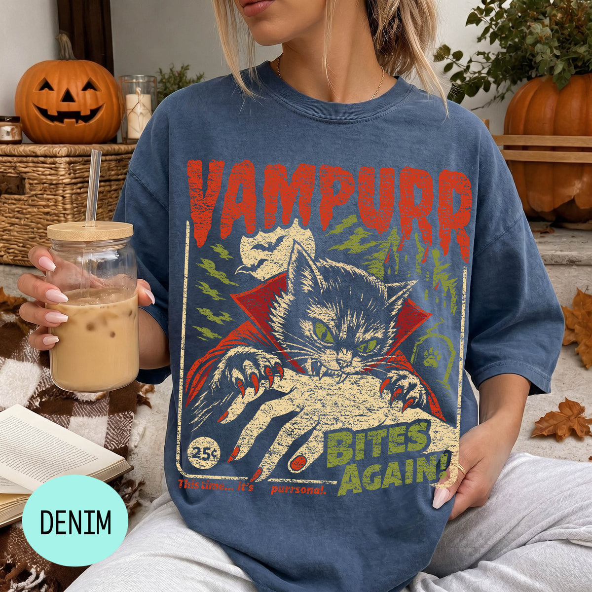 A woman wearing a blue t-shirt with a graphic design of a cat and the text "VAMPURR BITE'S AGAIN!" is sitting on a couch with a pumpkin and a drink in her hand.