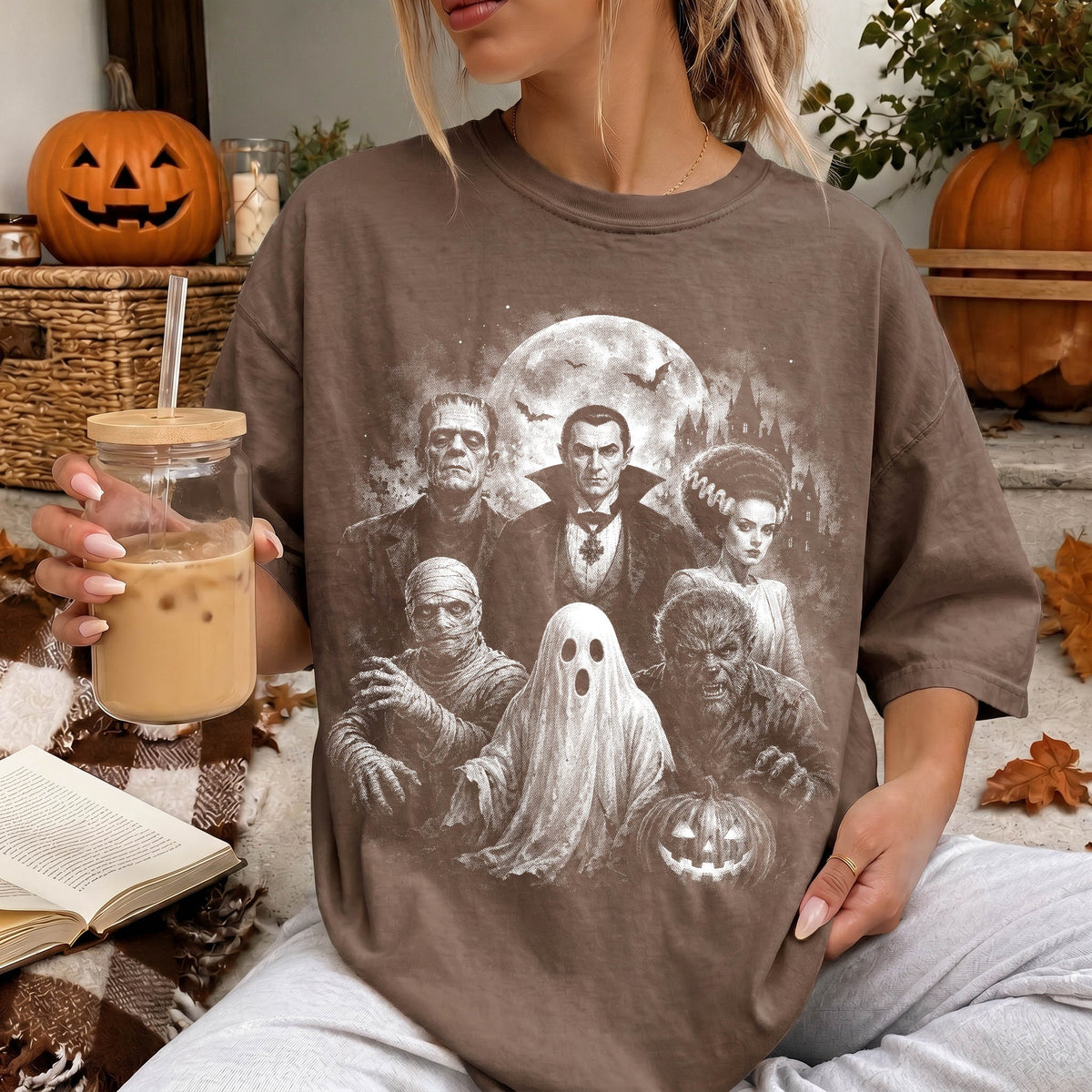 A woman is sitting on a couch, wearing a t-shirt with a Halloween-themed graphic design featuring characters such as Dracula, Frankenstein, and a ghost. She is holding a glass of iced coffee and has a book nearby.