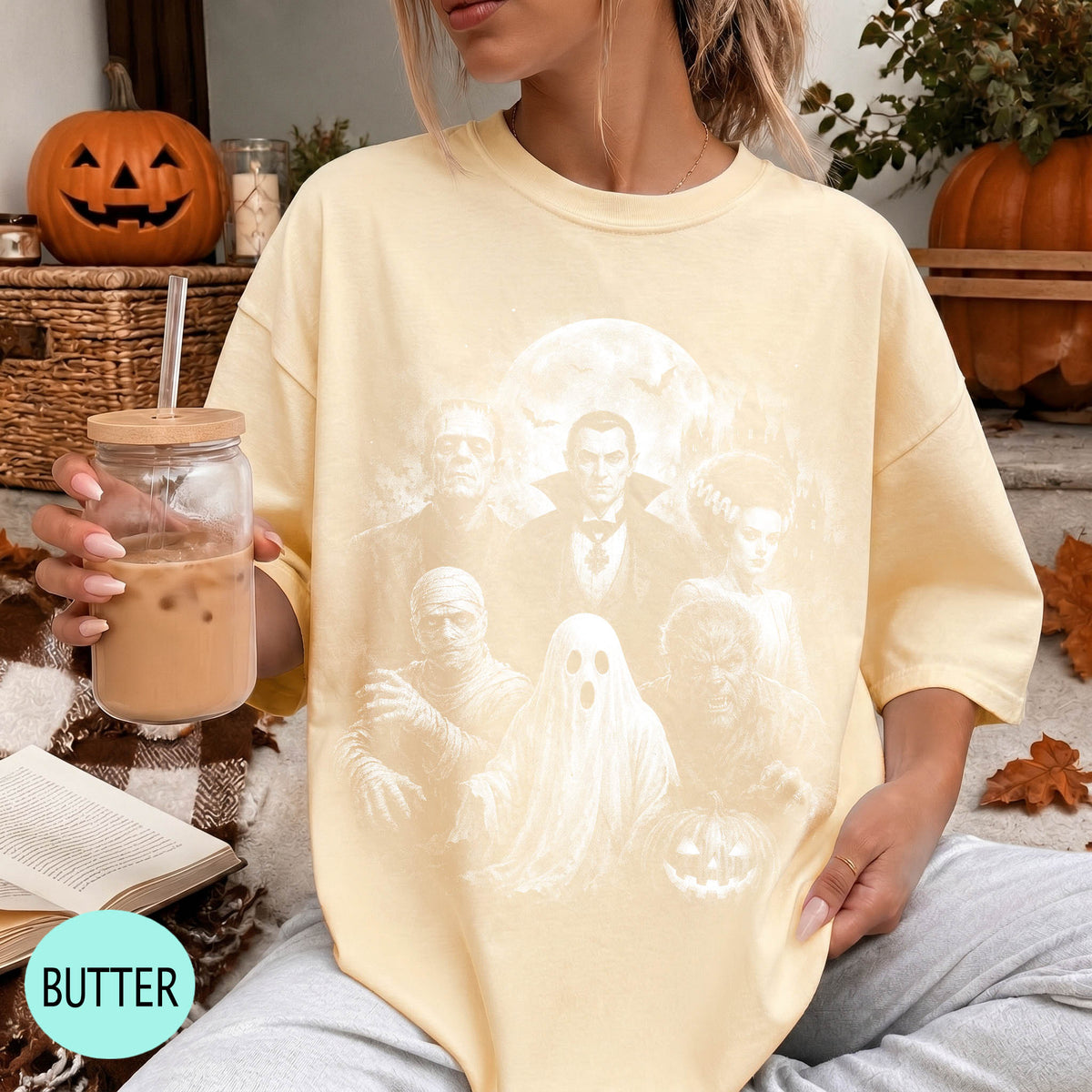 A woman wearing a yellow t-shirt with a Halloween-themed graphic, holding a drink and sitting on a blanket with a pumpkin and other fall decorations in the background.