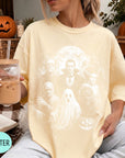 A woman wearing a yellow t-shirt with a Halloween-themed graphic, holding a drink and sitting on a blanket with a pumpkin and other fall decorations in the background.