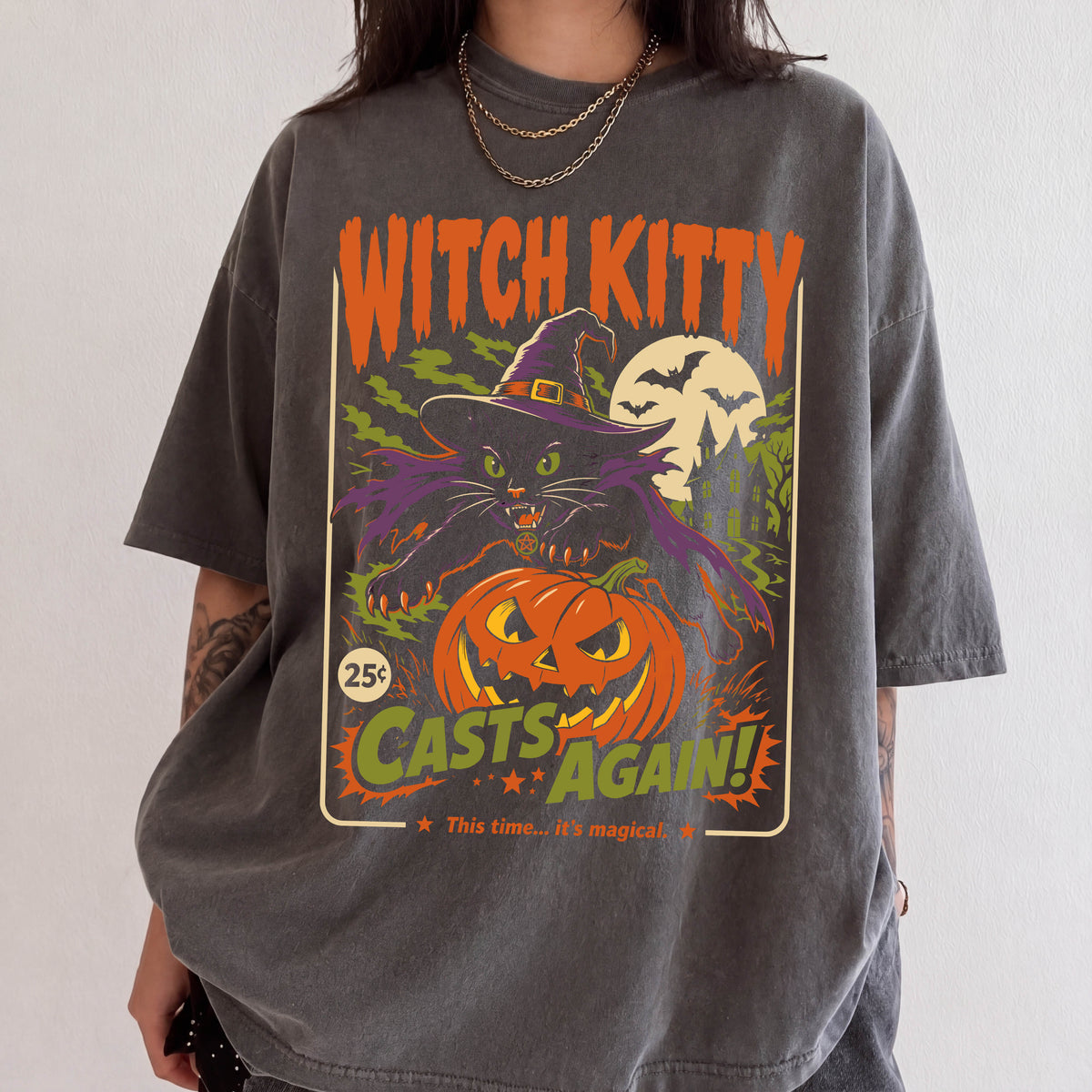 A person wearing a gray t-shirt with a Halloween-themed graphic design featuring a witch, a jack-o-lantern, and bats.