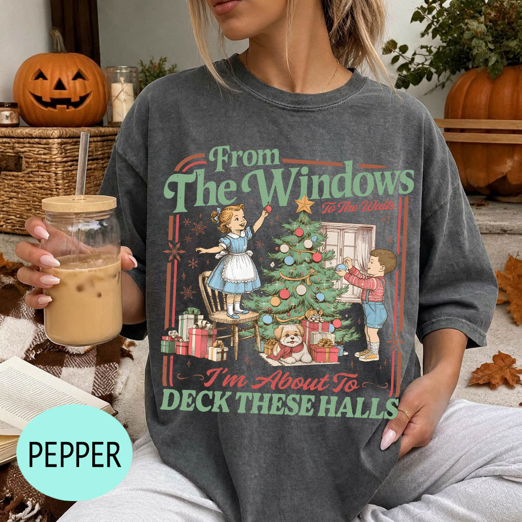 A woman wearing a gray t-shirt with a graphic design of a Christmas tree and two children decorating it, holding a drink in a glass.