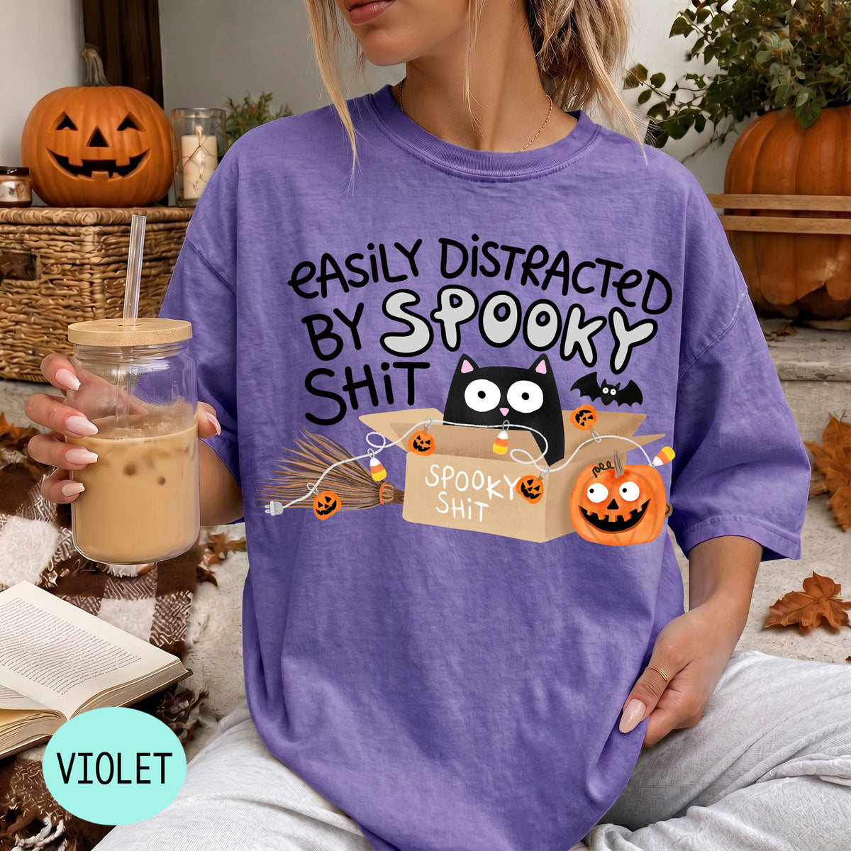 A person wearing a purple t-shirt with a humorous Halloween-themed graphic, including a cat, pumpkins, bats, and the text "EASILY DISTracted BY SPOOKY SHIT".