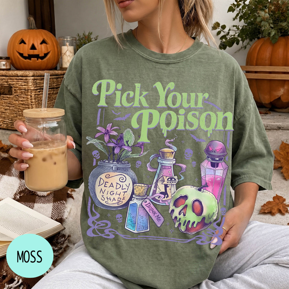 A woman wearing a green t-shirt with a Halloween-themed design, holding a drink and surrounded by Halloween decorations.