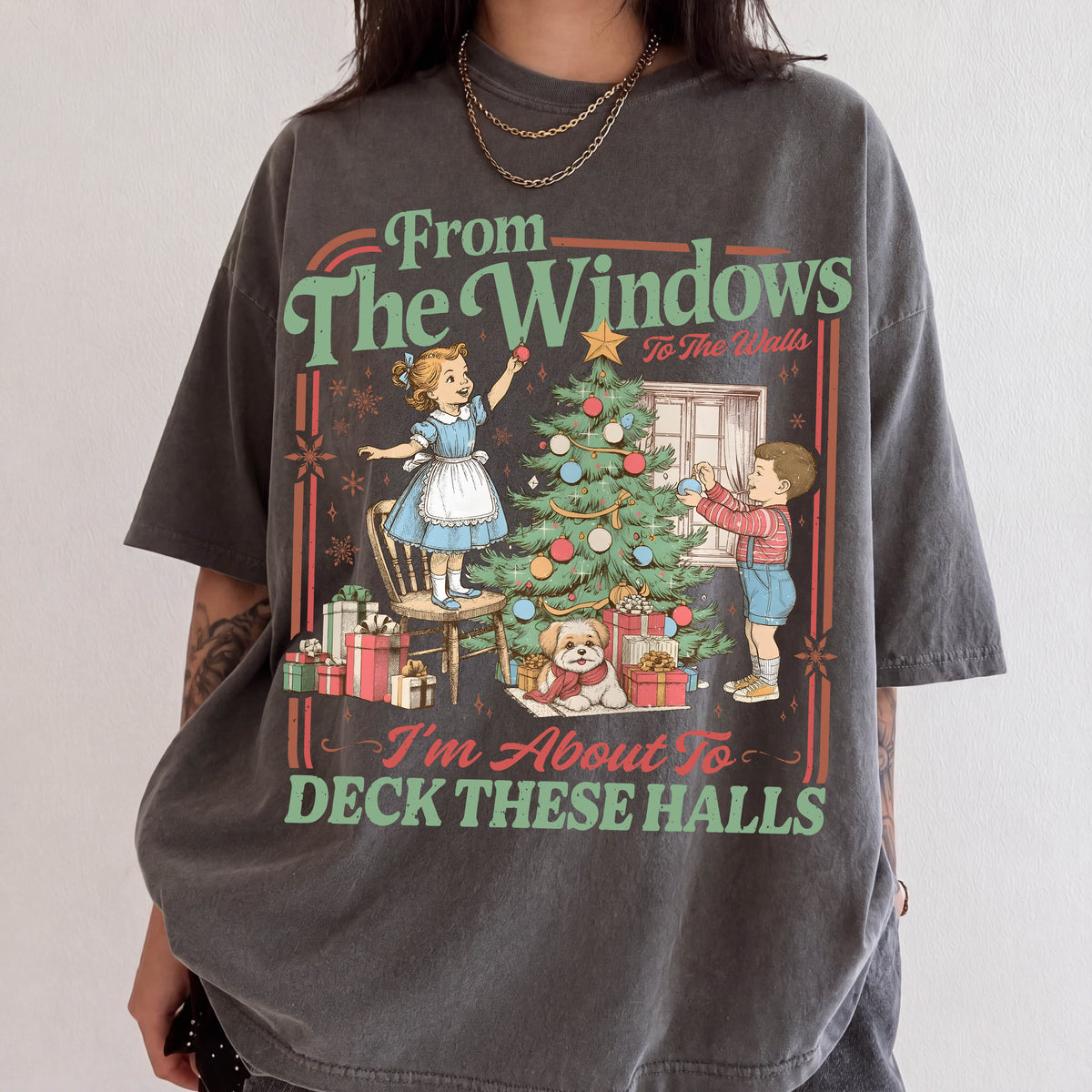 A person wearing a gray t-shirt with a Christmas-themed graphic design featuring a young girl decorating a Christmas tree and a boy holding a gift.