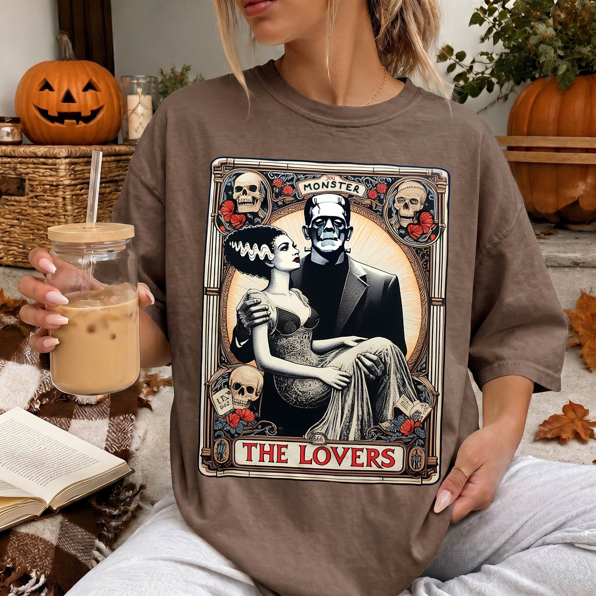 A woman wearing a t-shirt with a graphic design featuring a couple and the text "MONSTER THE LOVERS". She is holding a drink in her hand and there are Halloween decorations visible in the background.