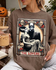 A woman wearing a t-shirt with a graphic design featuring a couple and the text "MONSTER THE LOVERS". She is holding a drink in her hand and there are Halloween decorations visible in the background.
