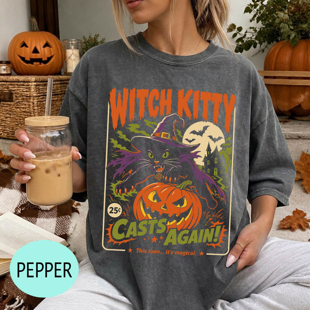 A woman wearing a gray t-shirt with a Halloween-themed graphic design, including a witch, pumpkin, and bats. She is holding a drink in a glass with a straw.