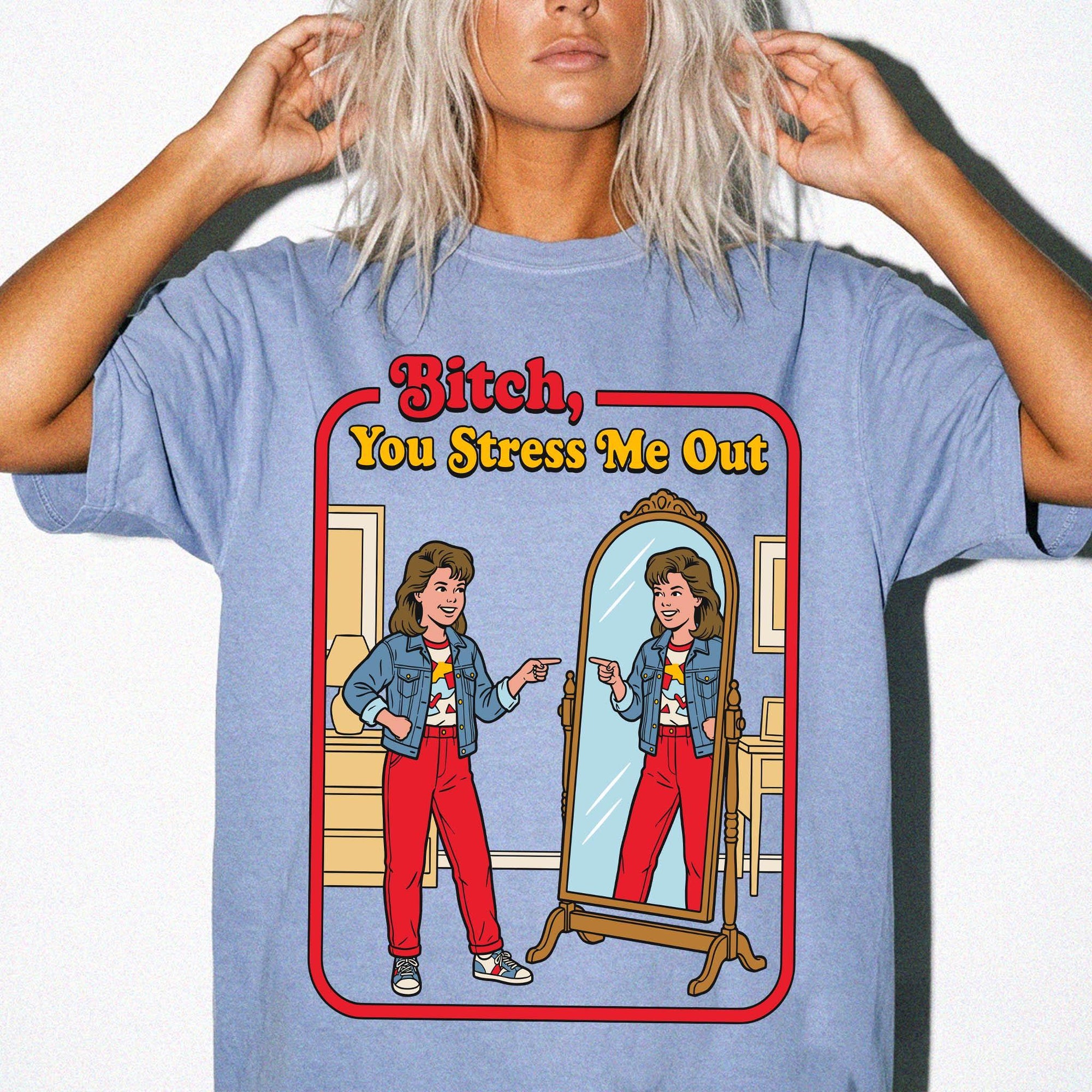 A woman with blonde hair wearing a blue t-shirt with a graphic design depicting a woman looking into a mirror and pointing at herself.