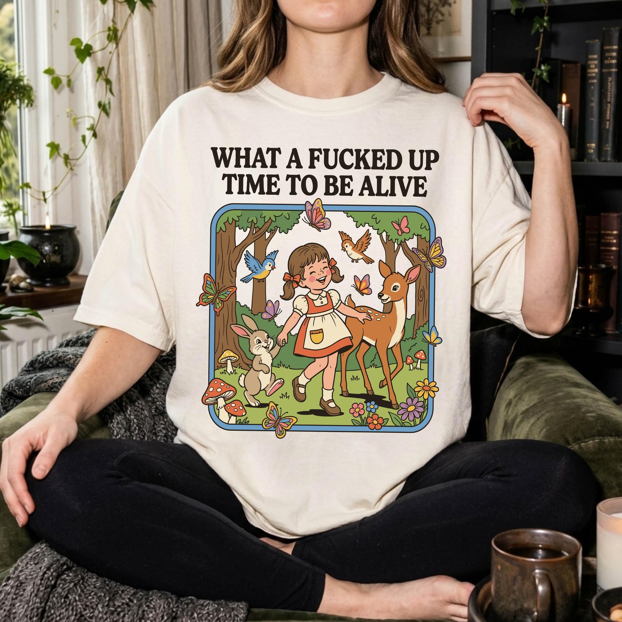 A person wearing a white t-shirt with a graphic design depicting a girl surrounded by various animals, including a deer, butterflies, and mushrooms, along with the text "What a FUCKED UP TIME TO BE ALIVE".