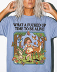 A woman wearing a blue t-shirt with a graphic design depicting a young girl surrounded by various animals, including a deer, rabbit, and butterflies, along with the text "WHAT A FUCKED UP TIME TO BE ALIVE".