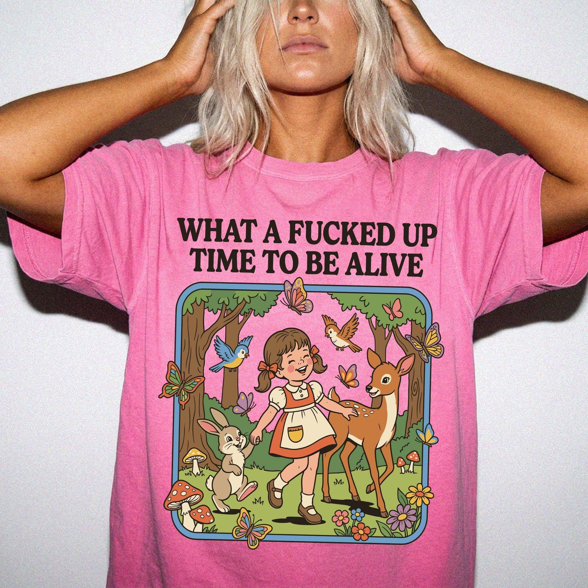 A woman wearing a pink t-shirt with a cartoon illustration of a girl interacting with a deer and other animals in a forest setting.