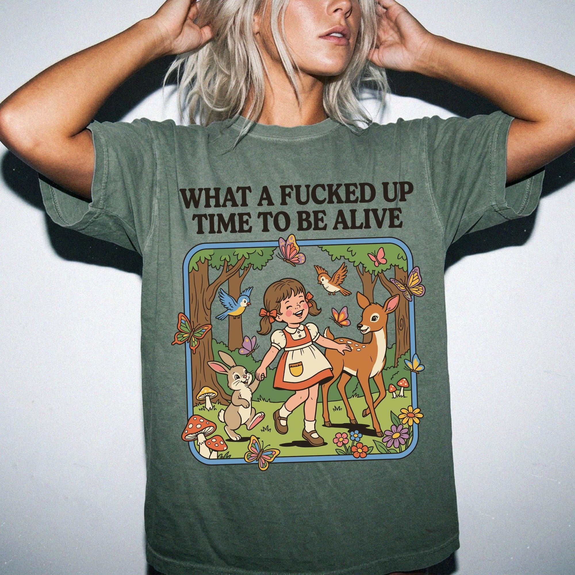 A woman wearing a green t-shirt with a cartoon illustration of a girl surrounded by animals and flowers, along with the text &amp;quot;What a FUCKED UP TIME TO BE ALIVE&amp;quot;.