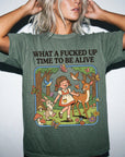 A woman wearing a green t-shirt with a cartoon illustration of a girl surrounded by animals and flowers, along with the text "What a FUCKED UP TIME TO BE ALIVE".