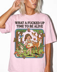 A woman wearing a pink t-shirt with a cartoon illustration of a girl surrounded by animals and flowers, along with the text "What a FUCKED UP TIME TO BE ALIVE".