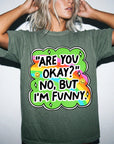 A person wearing a green t-shirt with a colorful graphic and text that says "ARE YOU OKAY? NO, BUT I&