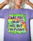 A person wearing a purple t-shirt with a colorful graphic and text that says "ARE YOU OKAY? NO, BUT I&