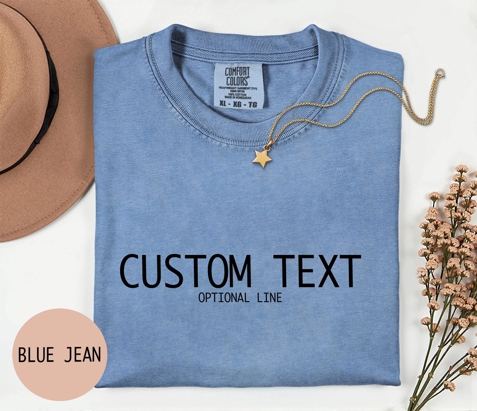 a blue denim t-shirt with the text &amp;quot;CUSTOM TEXT&amp;quot; printed on it, along with the phrase &amp;quot;OPTIONAL LINE&amp;quot; below it. The t-shirt is laid out on a white surface, accompanied by a beige hat, a gold chain, and some pink flowers.